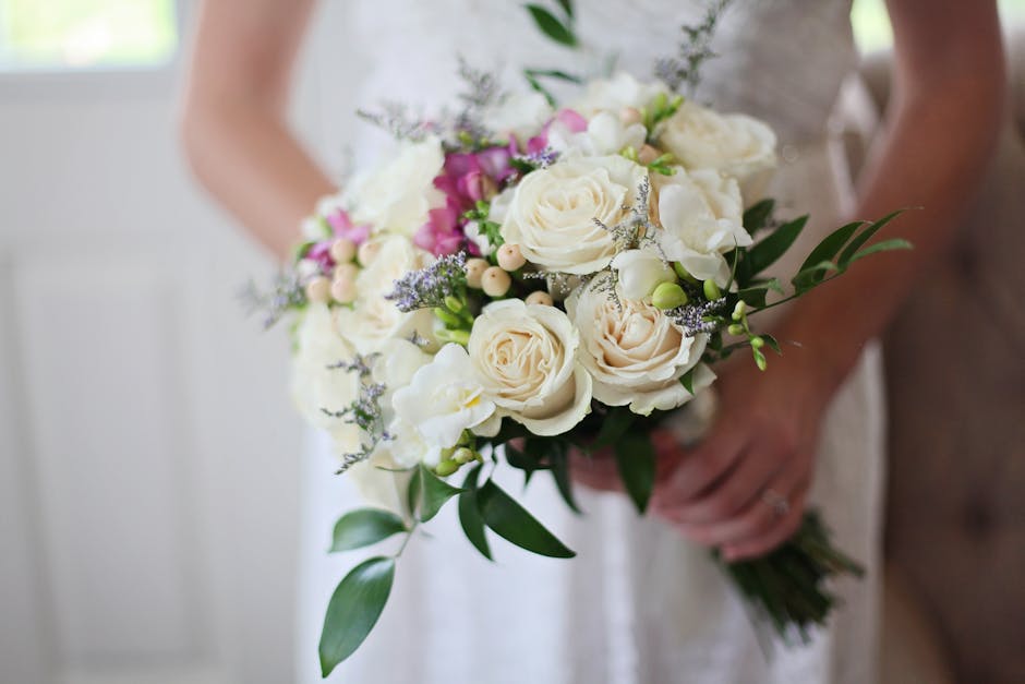 A close-up of a bridal bouquet held by a person in a white dress, featuring a carefully arranged assortment of fresh cream and blush roses, complemented by small pink and white buds, delicate lavender