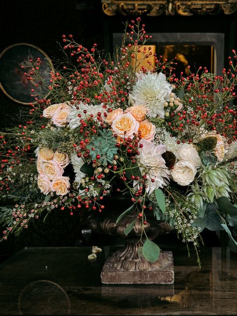 A lush floral arrangement displayed on a dark wooden table, featuring a mix of cream and peach roses, white dahlias, and green foliage, accented with red berry sprigs and textured greenery. The bouque