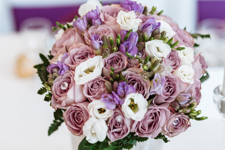 A close-up of a floral bouquet arranged with light pink roses, white lisianthus, and purple freesias, accented by small green leaves and a few decorative clear rhinestones. The flowers are fresh, with