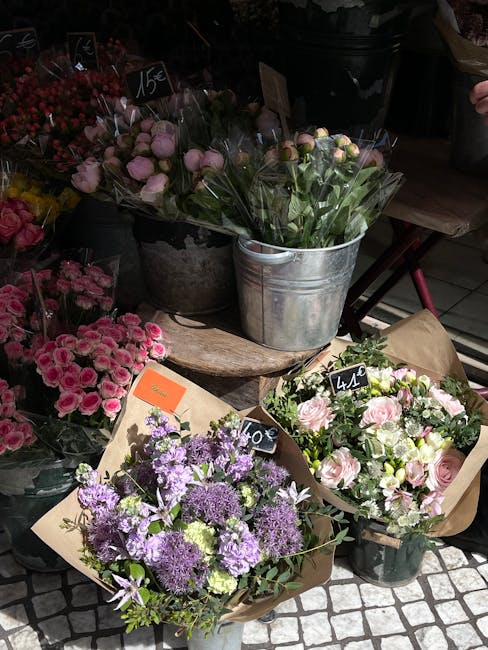 A display of fresh floral bouquets arranged in various containers at a flower shop, featuring a mix of pink roses, purple alliums, and pale pink ranunculus, wrapped in brown craft paper and placed in