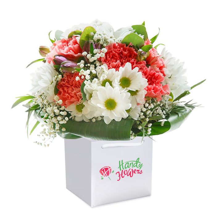 Balanced atop a clean, square white gift bag, this exuberant arrangement showcases a harmonious interaction of colours-peach and coral carnations blending seamlessly with snowy white daisies that feature vivid lime centres. The carnations, renowned for their plush, frilled petals, announce sheer abundance and texture, creating an interior warmth reminiscent of Bloomsbury's well-loved gardens. Dotted throughout the bouquet are delicate, cloud-like tufts of white baby's breath, providing an airy backdrop that lets every flower breathe, akin to soft spring breezes moving through Gordon Square. Subtle touches of unopened, lavender-highlighted buds peek from the left side, introducing a sense of mystery and gentle anticipation. Enveloping the flowers, wide and darkly lustrous leaves provide organic structure and a fresh undercurrent, supporting the blooms as gracefully as the ornate railings protect the area's historic squares. The bouquet's shape is invitingly round and robust, curated by a local florist who understands both the traditions and contemporary tastes of Bloomsbury. The crisp bag with its sturdy ribbon handles is not just practical for hand-delivery across WC1, but also sets the composition apart as a ready-to-gift treasure. This luminous ensemble is perfect for birthdays, anniversaries, or those spontaneous acts of kindness that mark life in central London-its freshness and beauty a subtle celebration of local artistry.